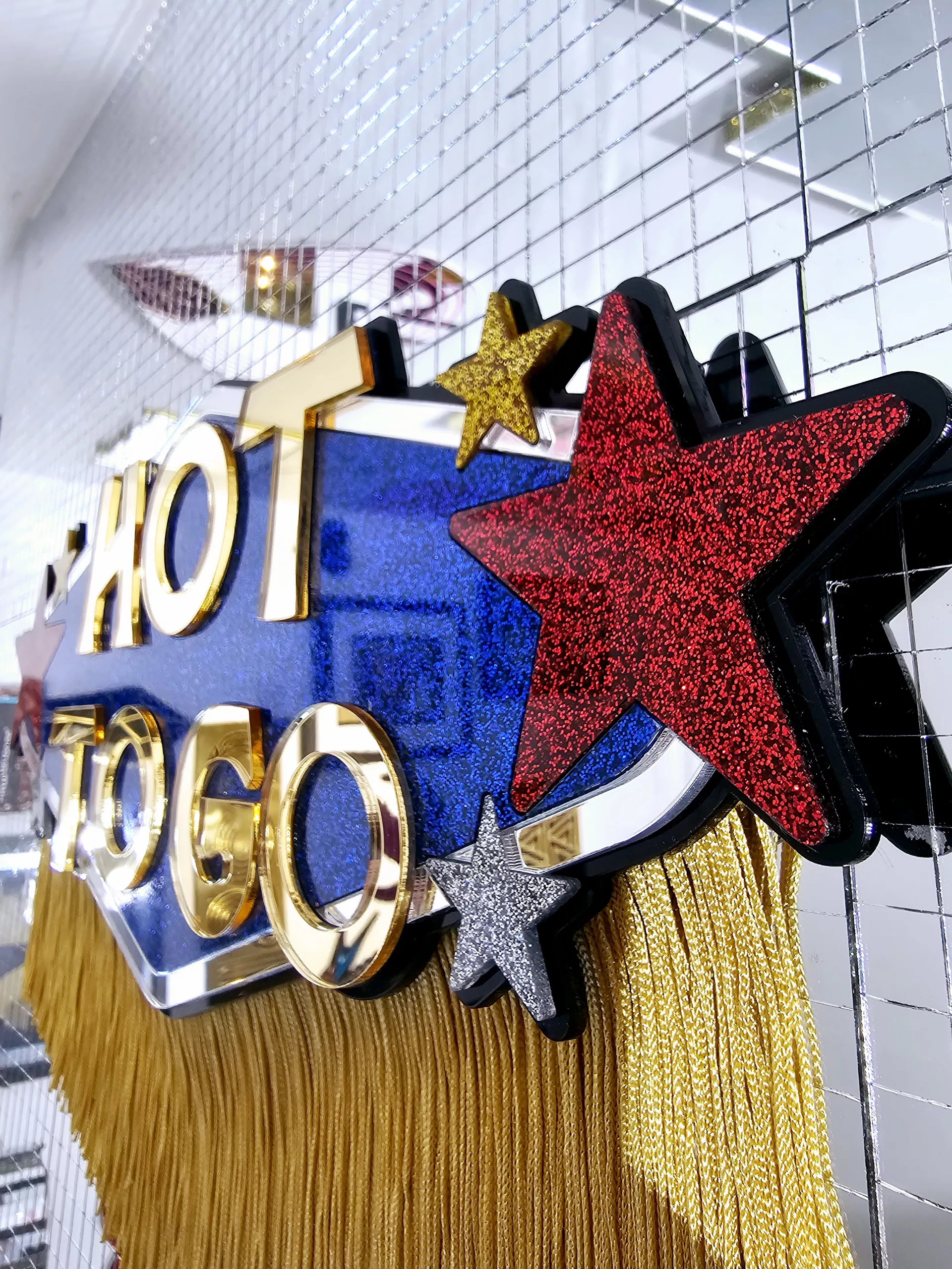 A piece of handmade wall art inspired by Chappell Roan's Hot To Go video. The piece is made with blue glitter, flanked with red, gold and silver glitter stars with the words Hot To Go in gold mirror, all finished with gold fringing.