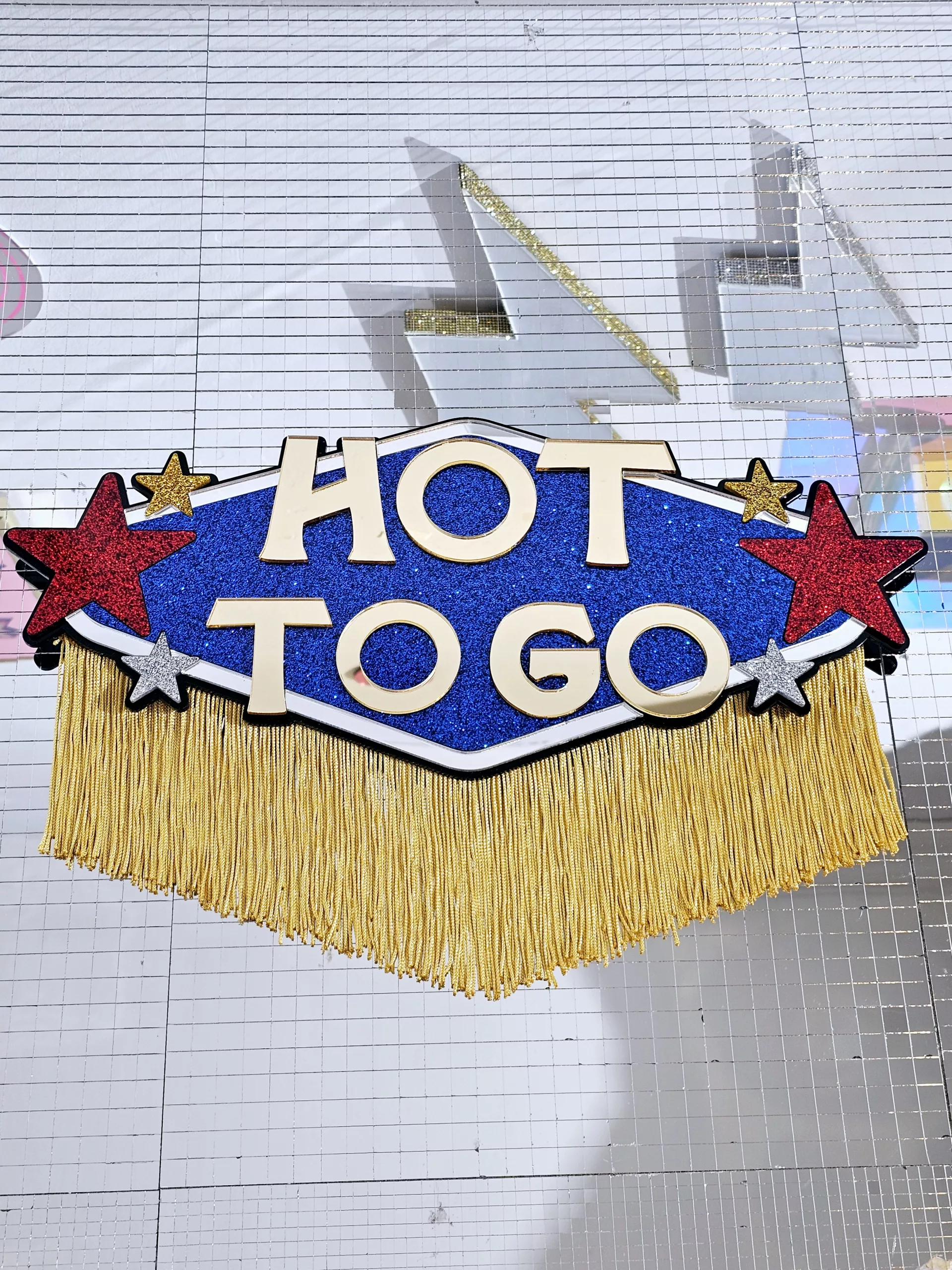 A piece of handmade wall art inspired by Chappell Roan's Hot To Go video. The piece is made with blue glitter, flanked with red, gold and silver glitter stars with the words Hot To Go in gold mirror, all finished with gold fringing.