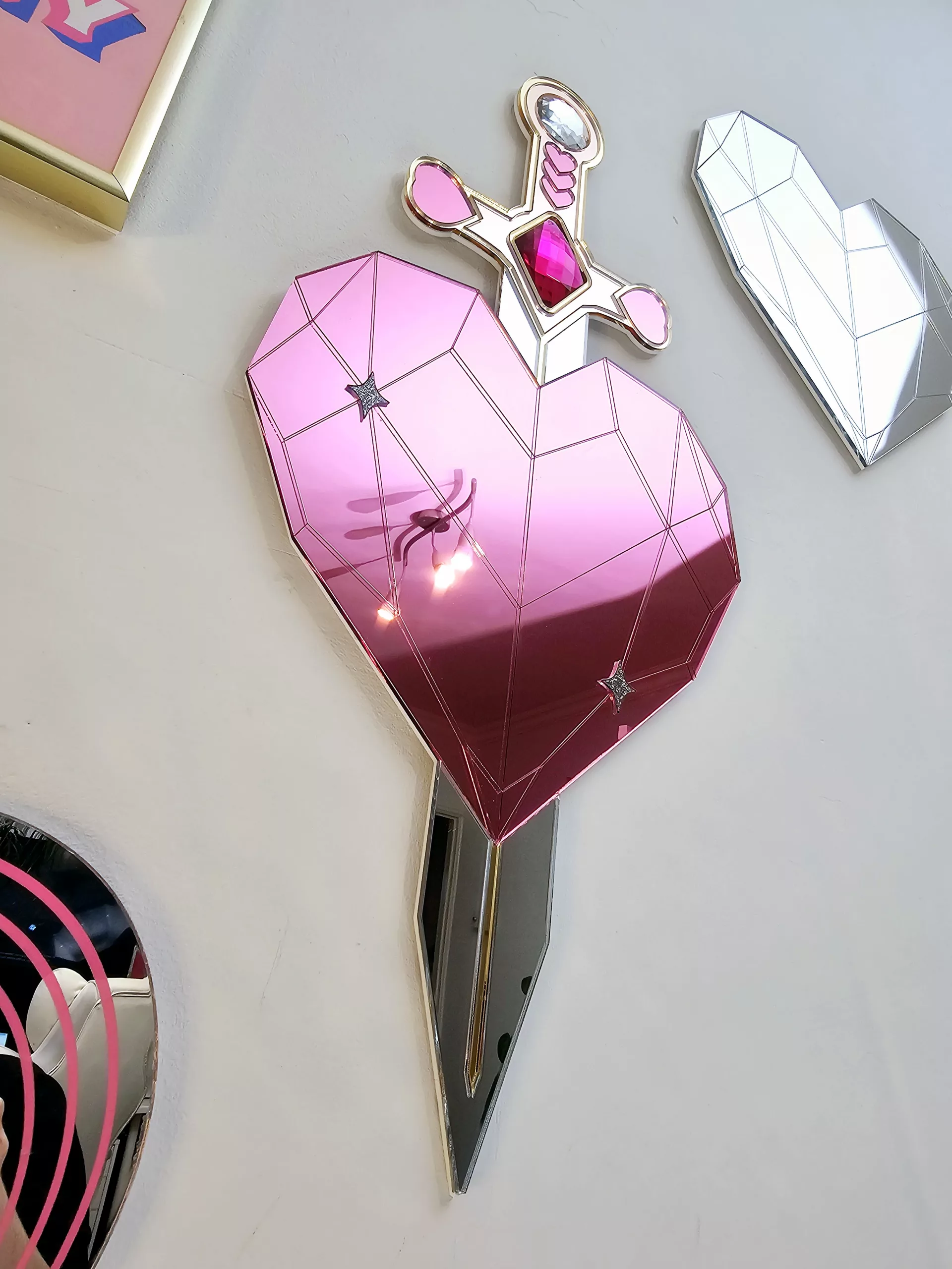A handmade mirror on the shape of a sword piercing a heart. Made with gold, rose gold, pink and silver mirror with gem accents.