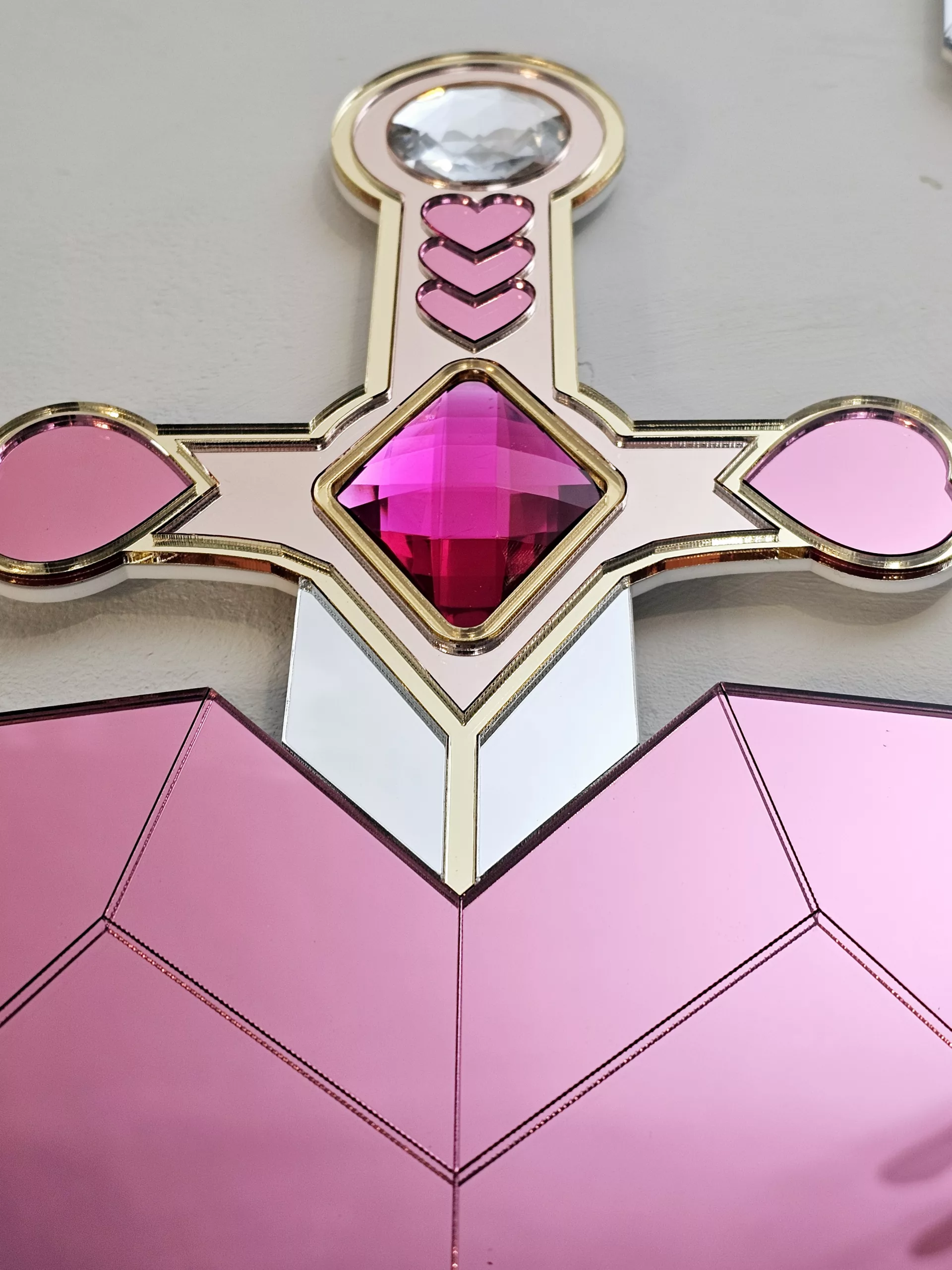 A handmade mirror on the shape of a sword piercing a heart. Made with gold, rose gold, pink and silver mirror with gem accents.