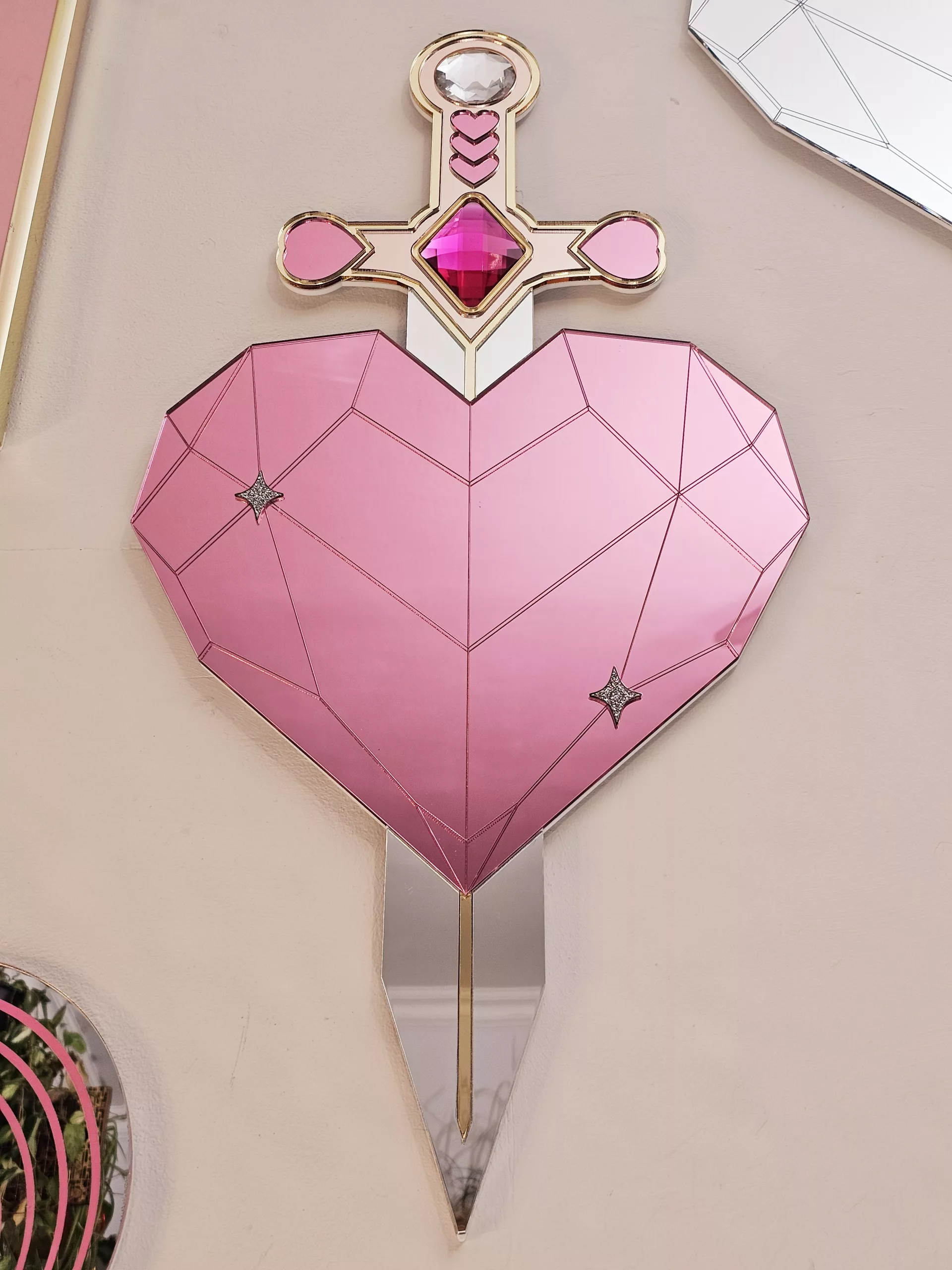 A handmade mirror on the shape of a sword piercing a heart. Made with gold, rose gold, pink and silver mirror with gem accents.