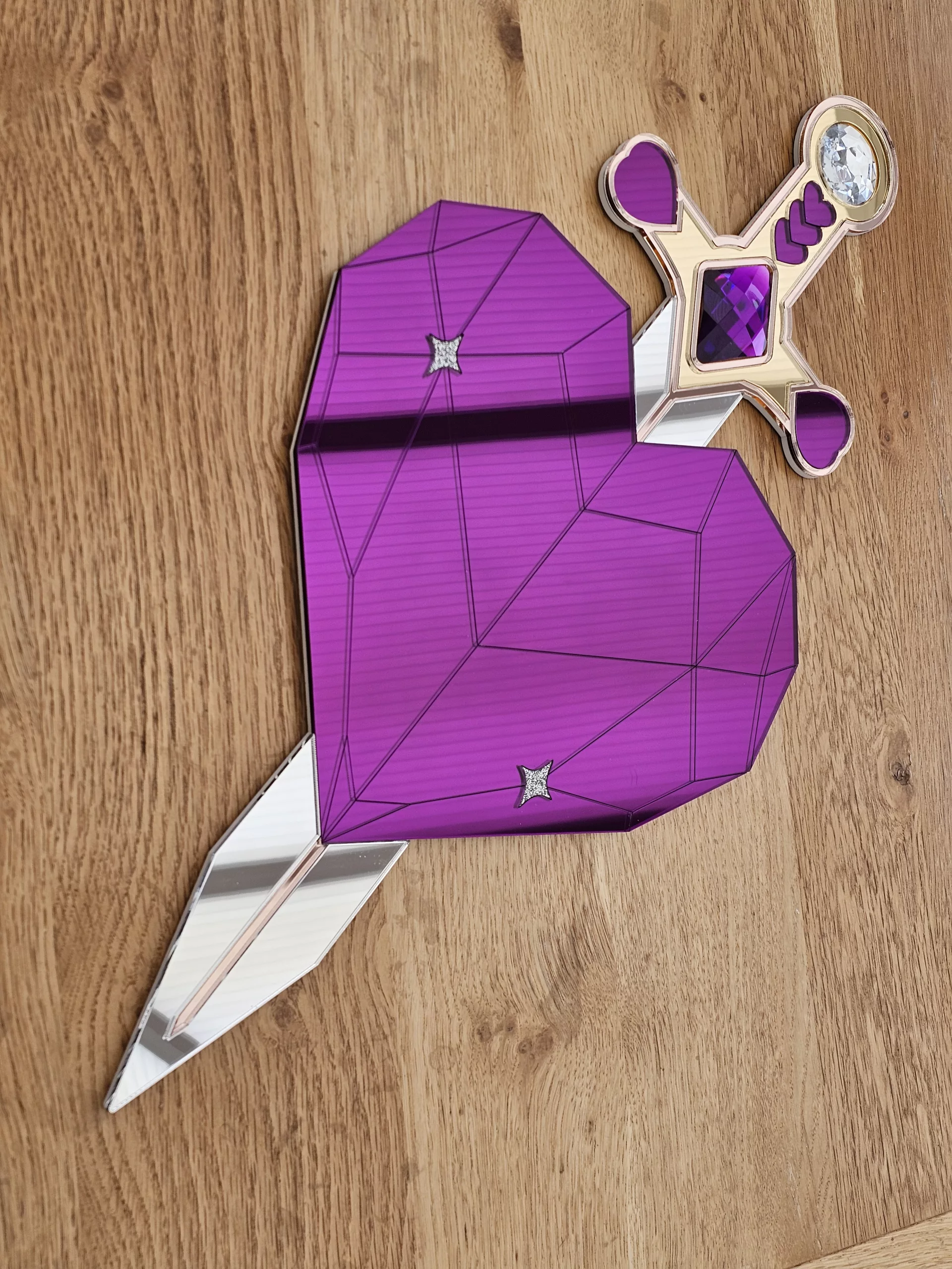 A handmade piece of wall art in the shape of a heart and sword. Made with purple mirror, gem and glitter accents.