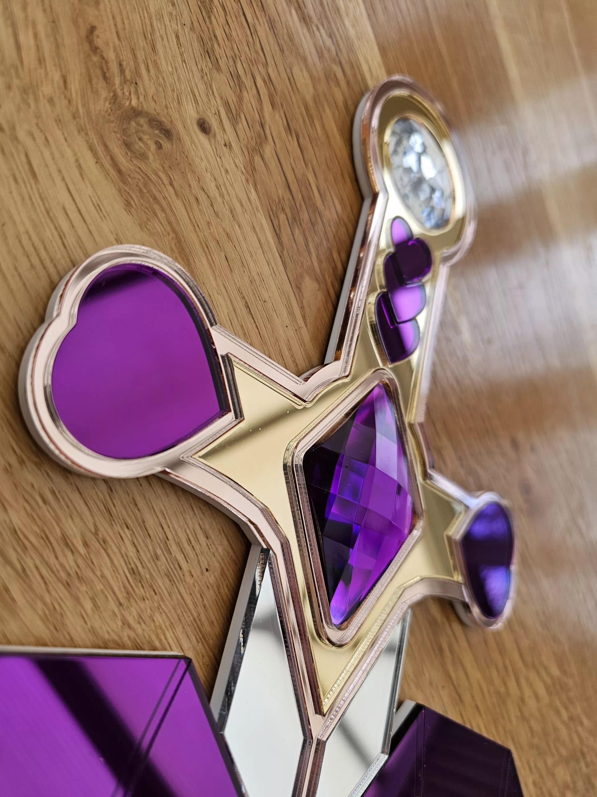 A handmade piece of wall art in the shape of a heart and sword. Made with purple mirror, gem and glitter accents.