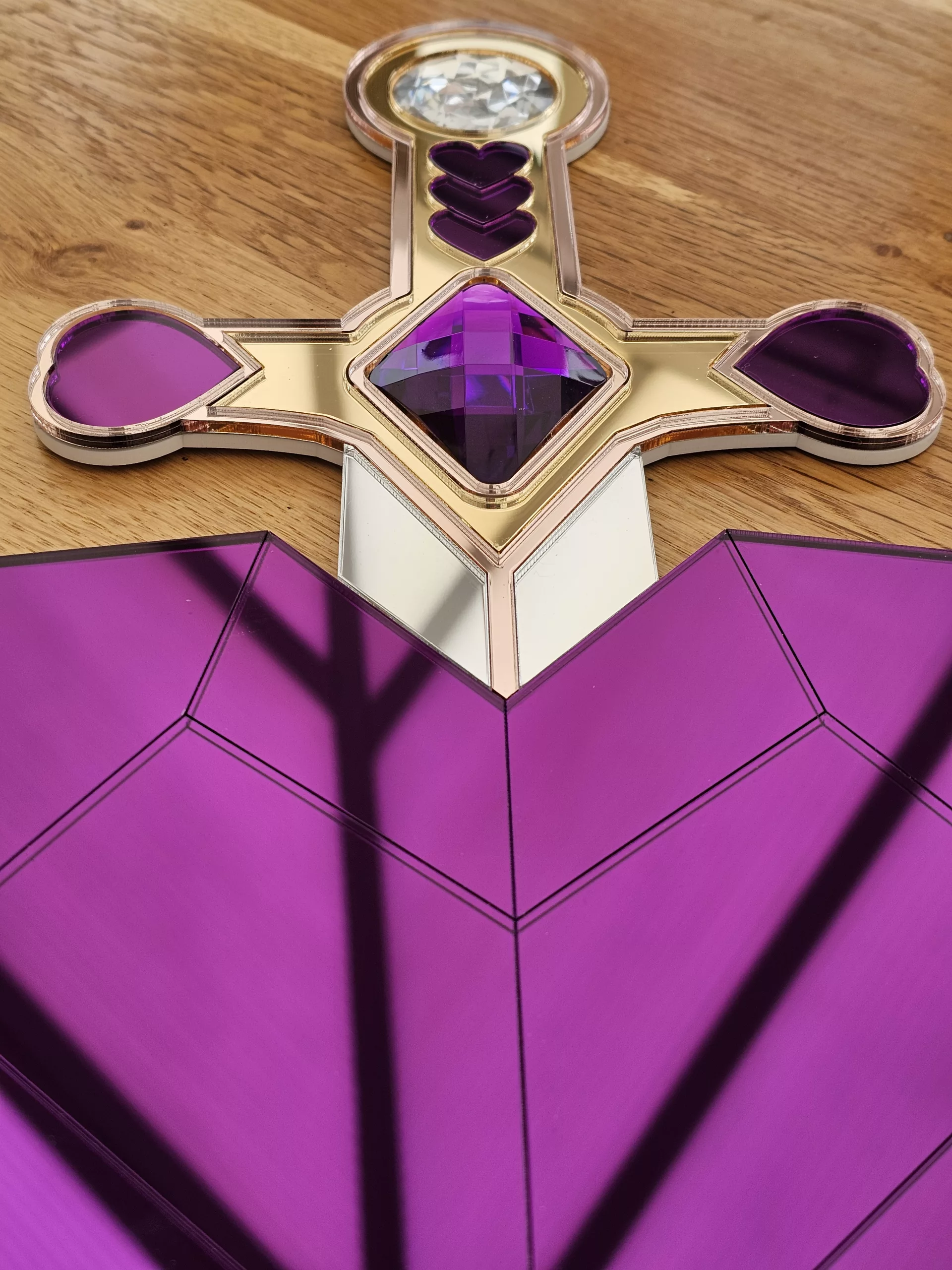 A handmade piece of wall art in the shape of a heart and sword. Made with purple mirror, gem and glitter accents.