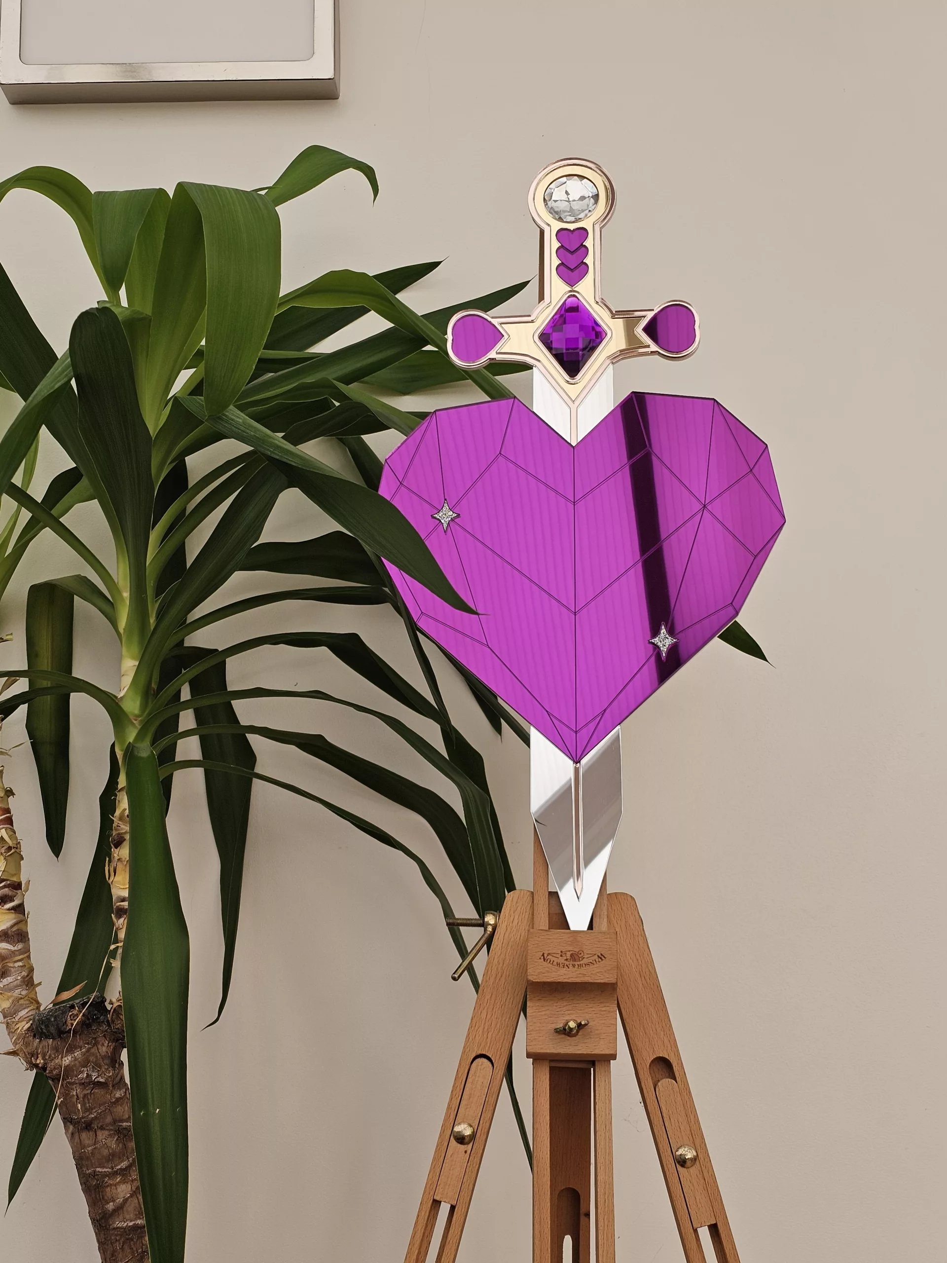 A handmade piece of wall art in the shape of a heart and sword. Made with purple mirror, gem and glitter accents.