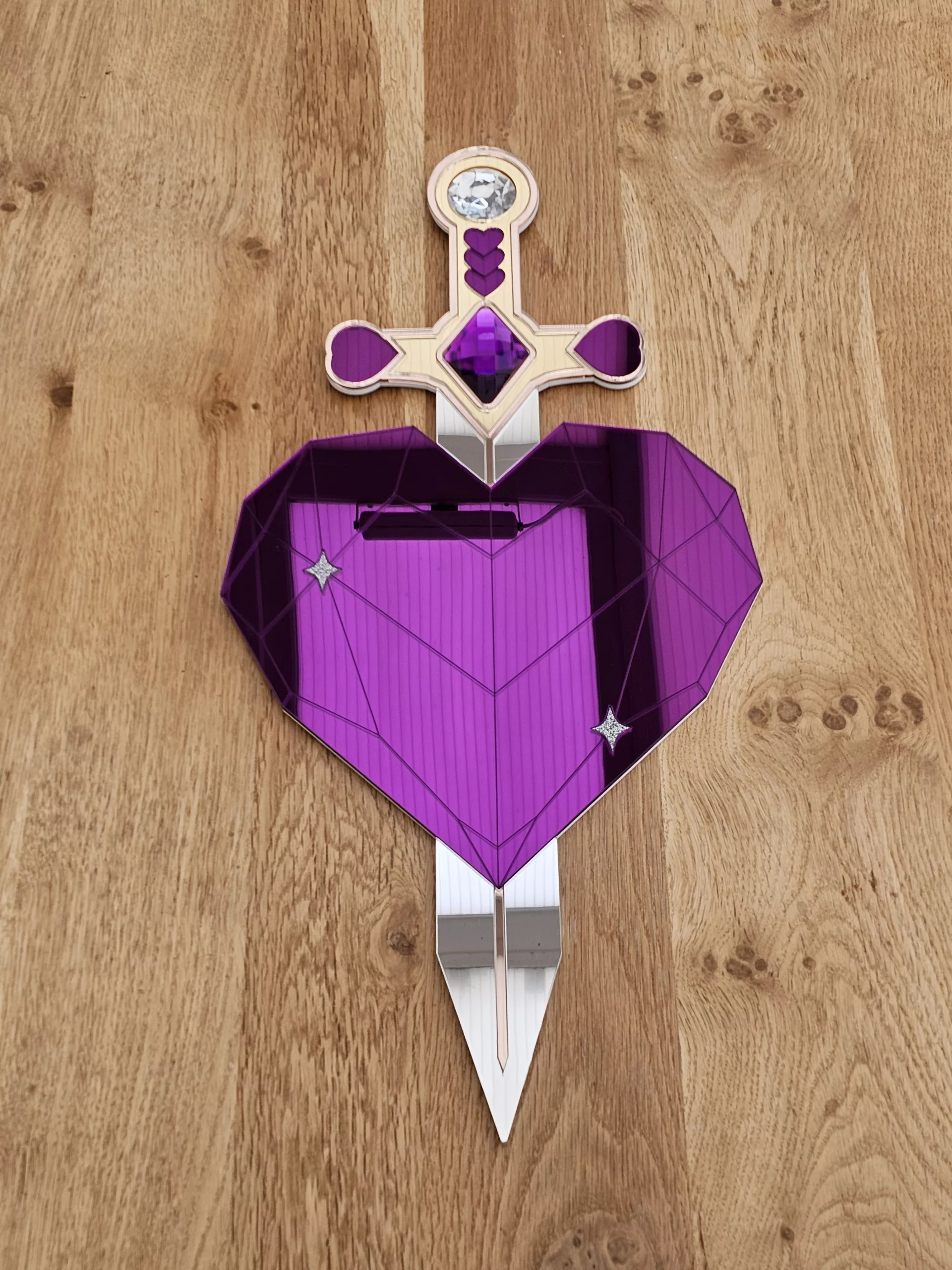A handmade piece of wall art in the shape of a heart and sword. Made with purple mirror, gem and glitter accents.
