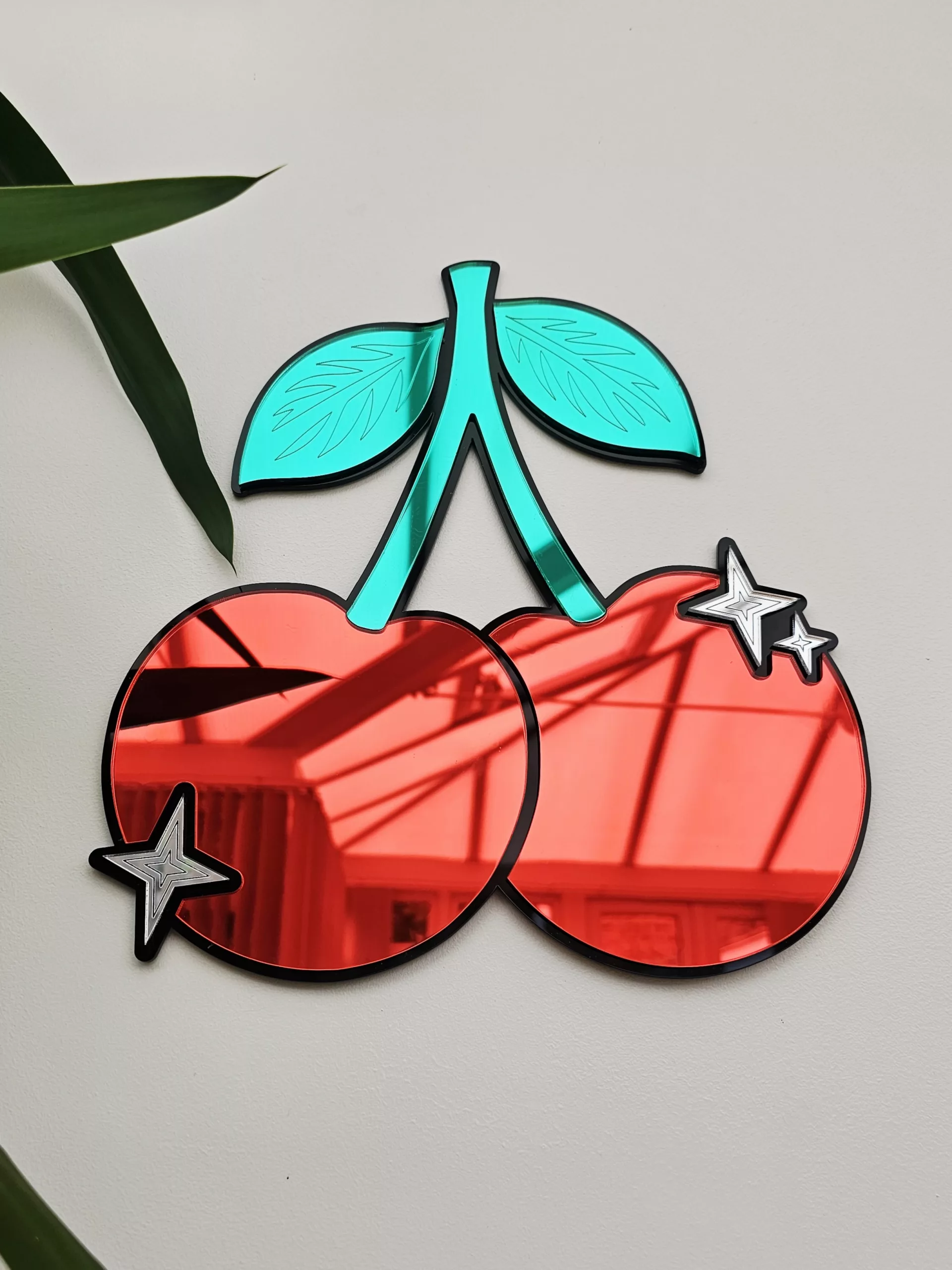 A piece of mirrored wall art in the shape of a cherry. With green leaves and silver sparkle details.