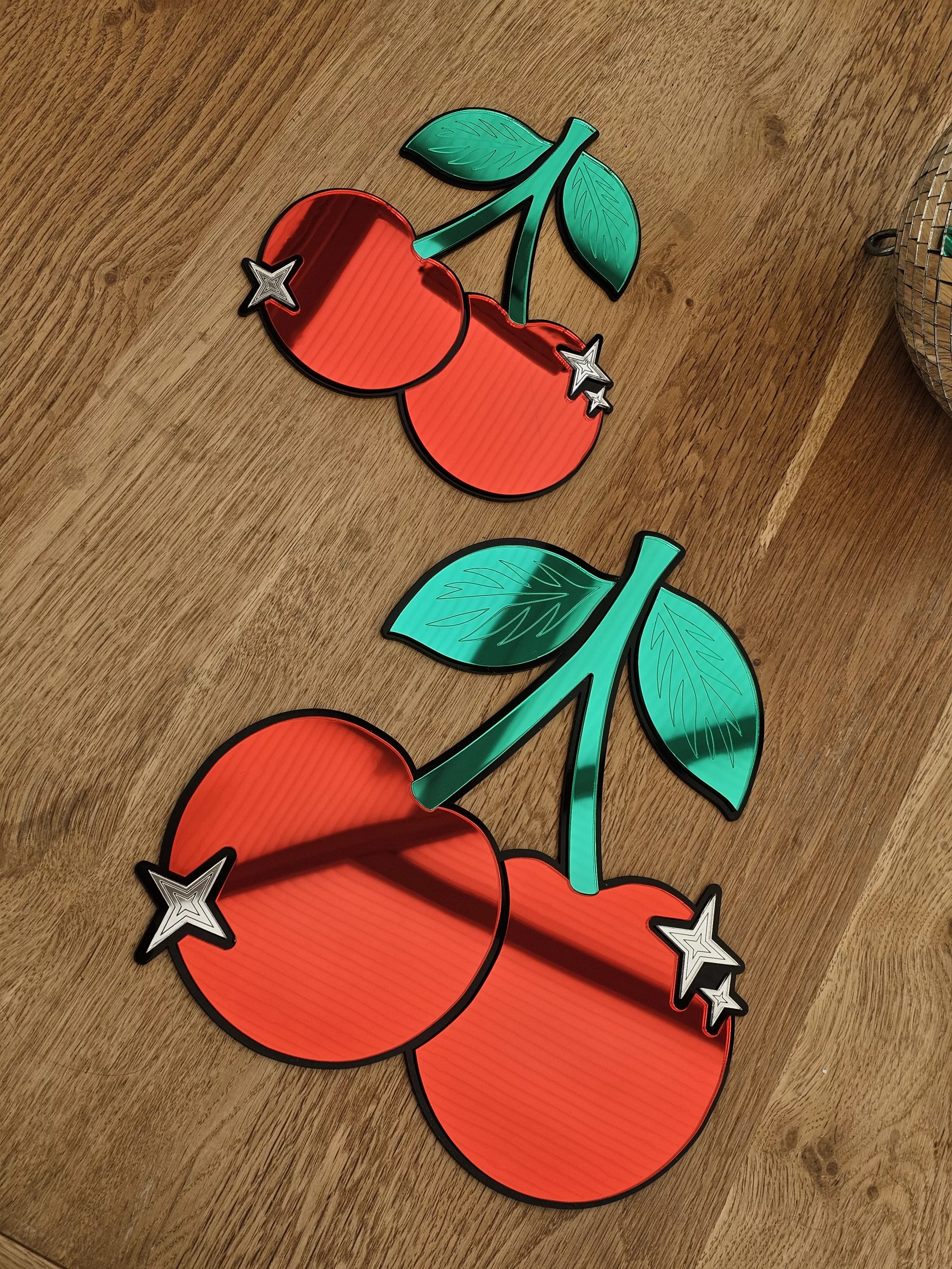 A piece of mirrored wall art in the shape of a cherry. With green leaves and silver sparkle details.