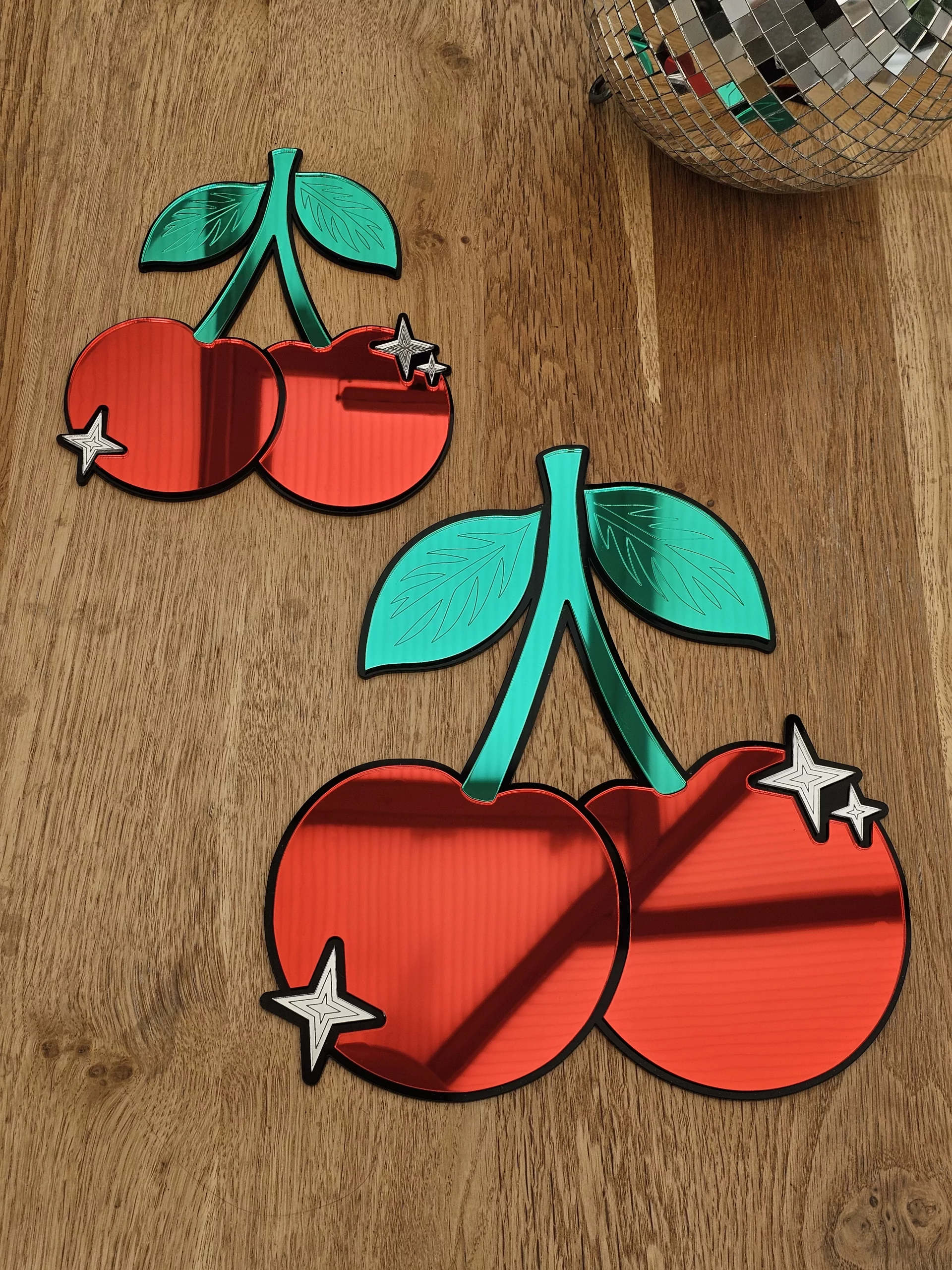 A piece of mirrored wall art in the shape of a cherry. With green leaves and silver sparkle details.