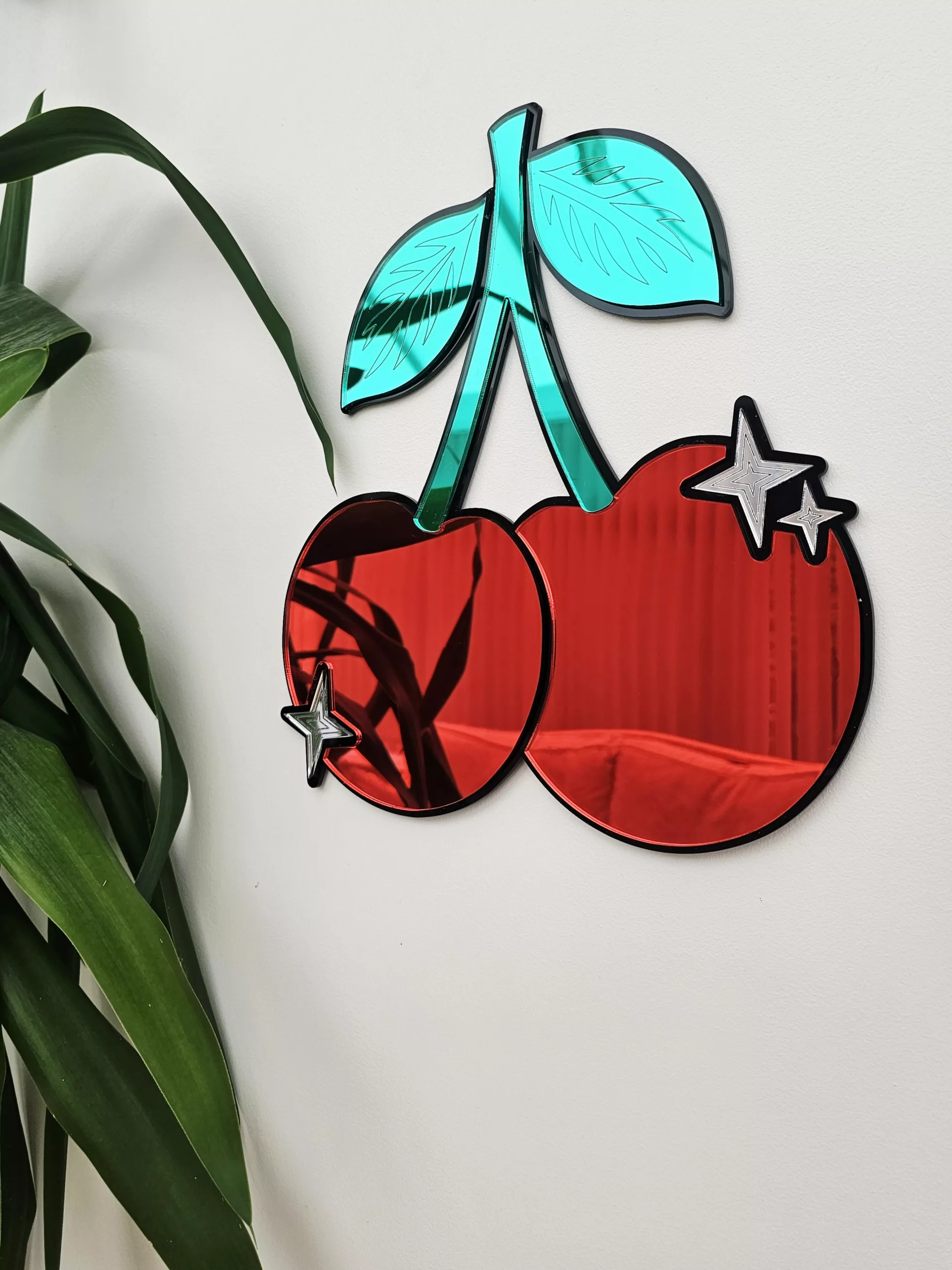 A piece of mirrored wall art in the shape of a cherry. With green leaves and silver sparkle details.
