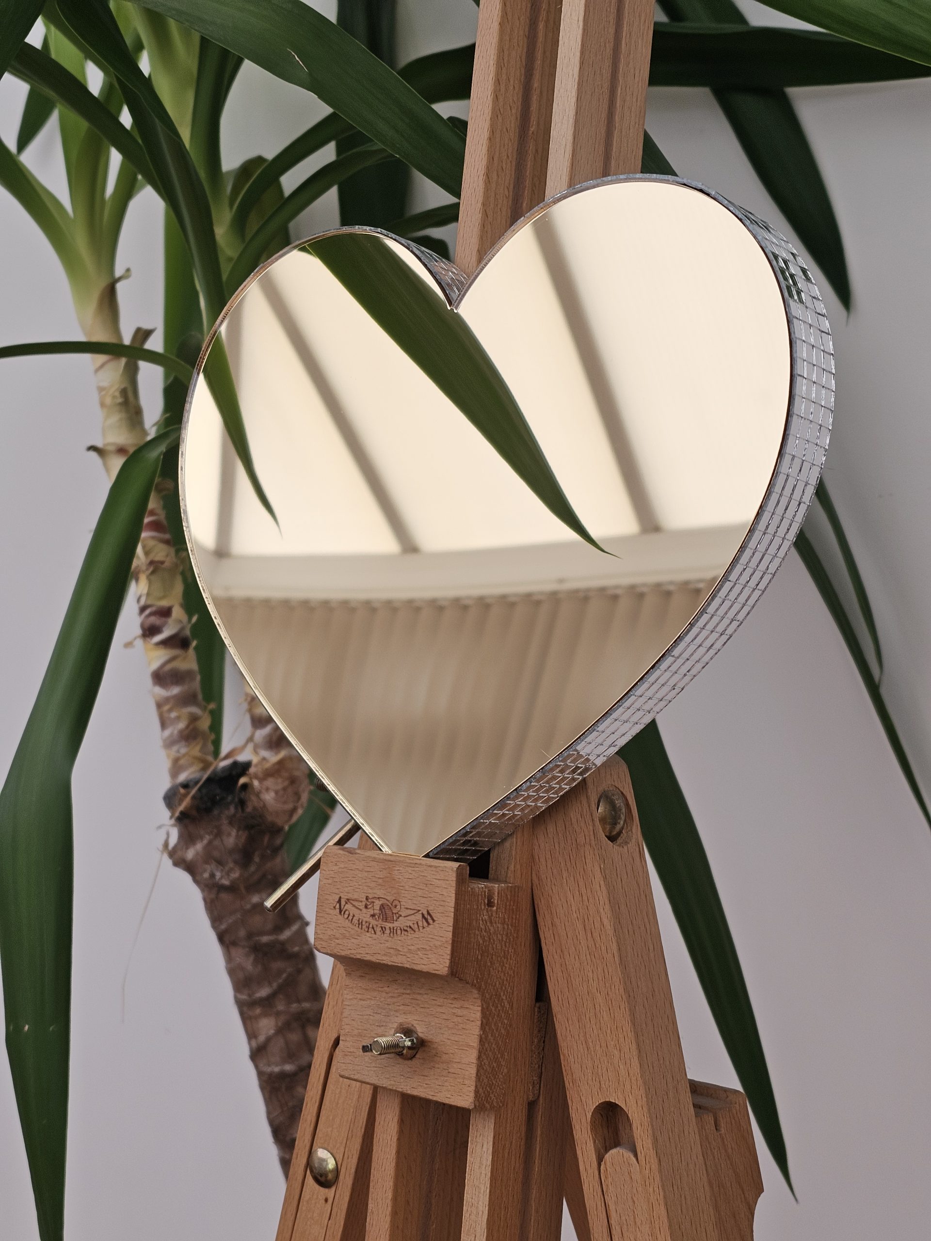 A gold mirror in the shape of a heart, with silver glass mirror tiles around the edges, perched on a wooden easel.