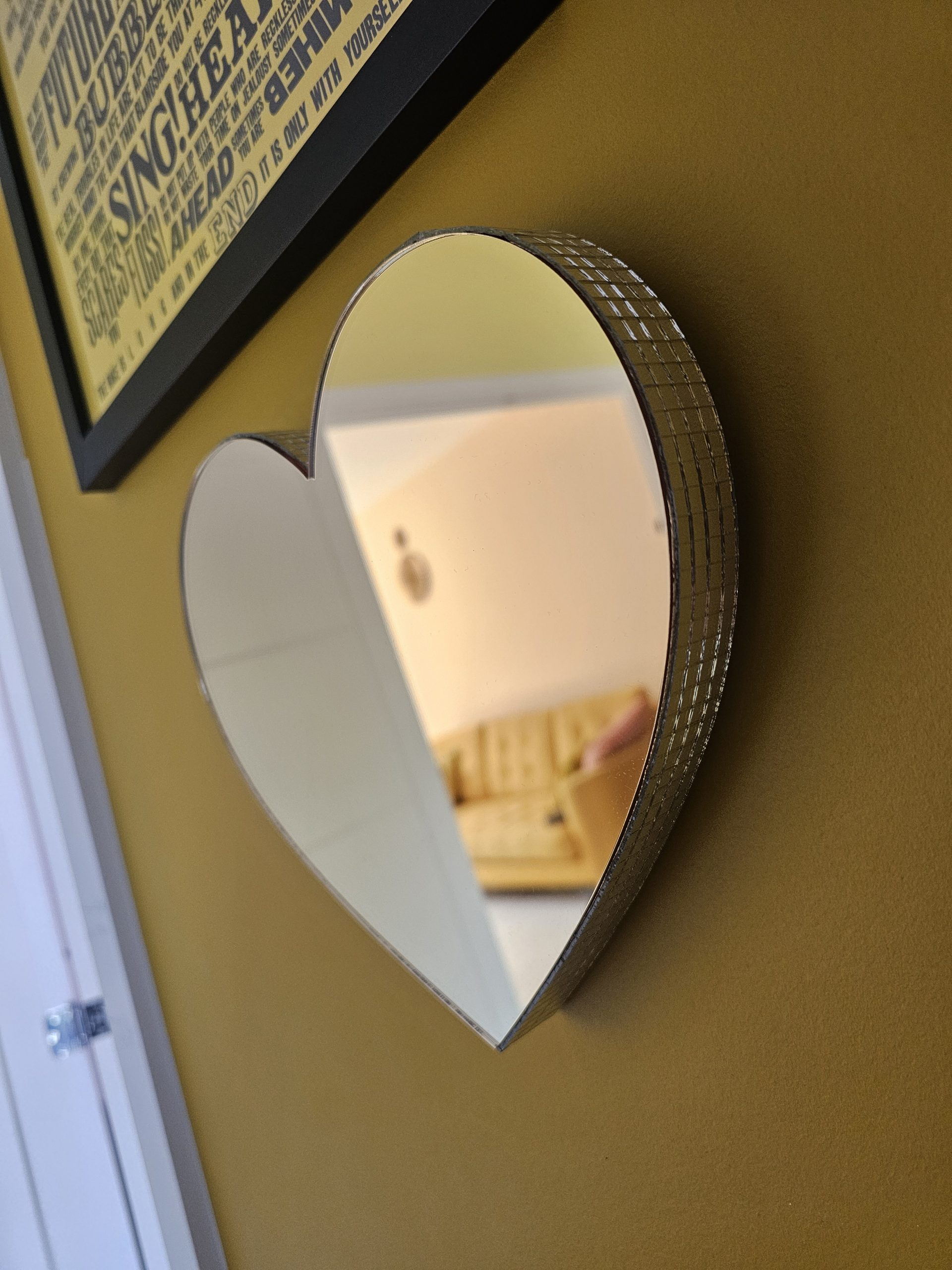 A gold mirror in the shape of a heart, with silver glass mirror tiles around the edges, mounted on an ochre coloured wall.