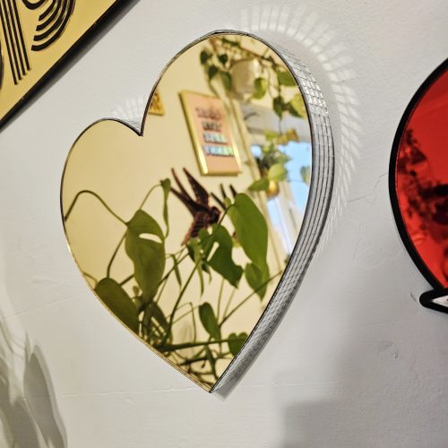 A gold mirror in the shape of a heart, with silver glass mirror tiles around the edges, mounted on a white wall.