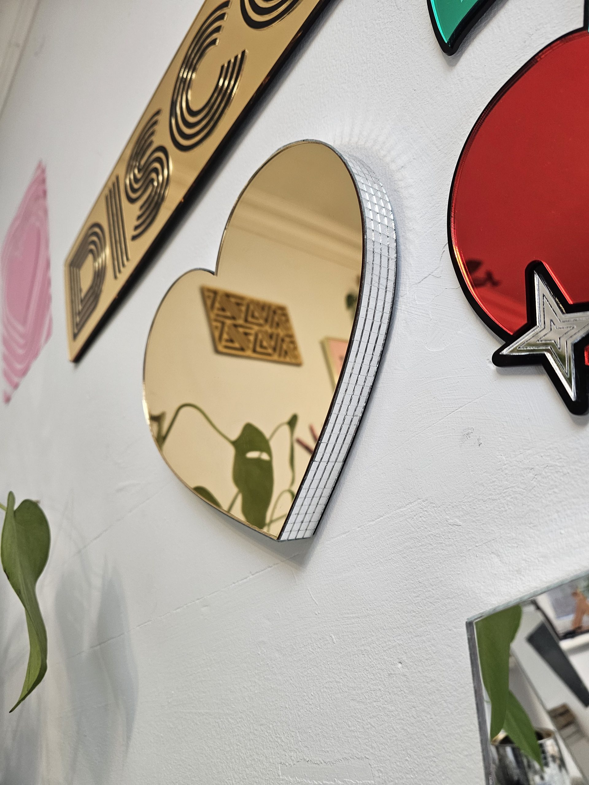 A gold mirror in the shape of a heart, with silver glass mirror tiles around the edges, mounted on a white wall.