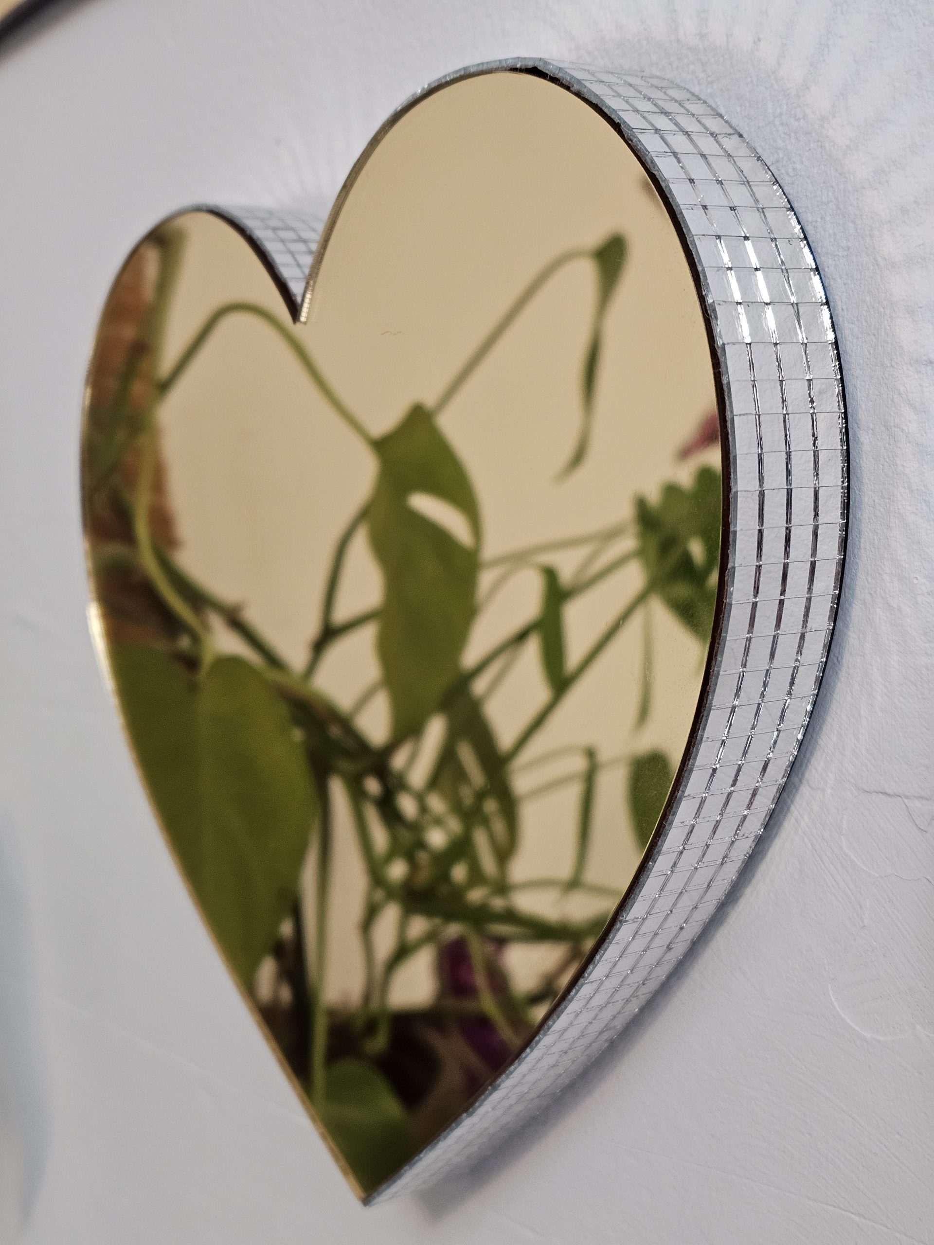 A gold mirror in the shape of a heart, with silver glass mirror tiles around the edges, mounted on a white wall.