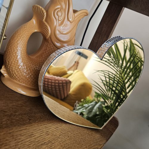 A gold mirror in the shape of a heart, with silver glass mirror tiles around the edges, perched on a shelf next to a vintage fish ornament.