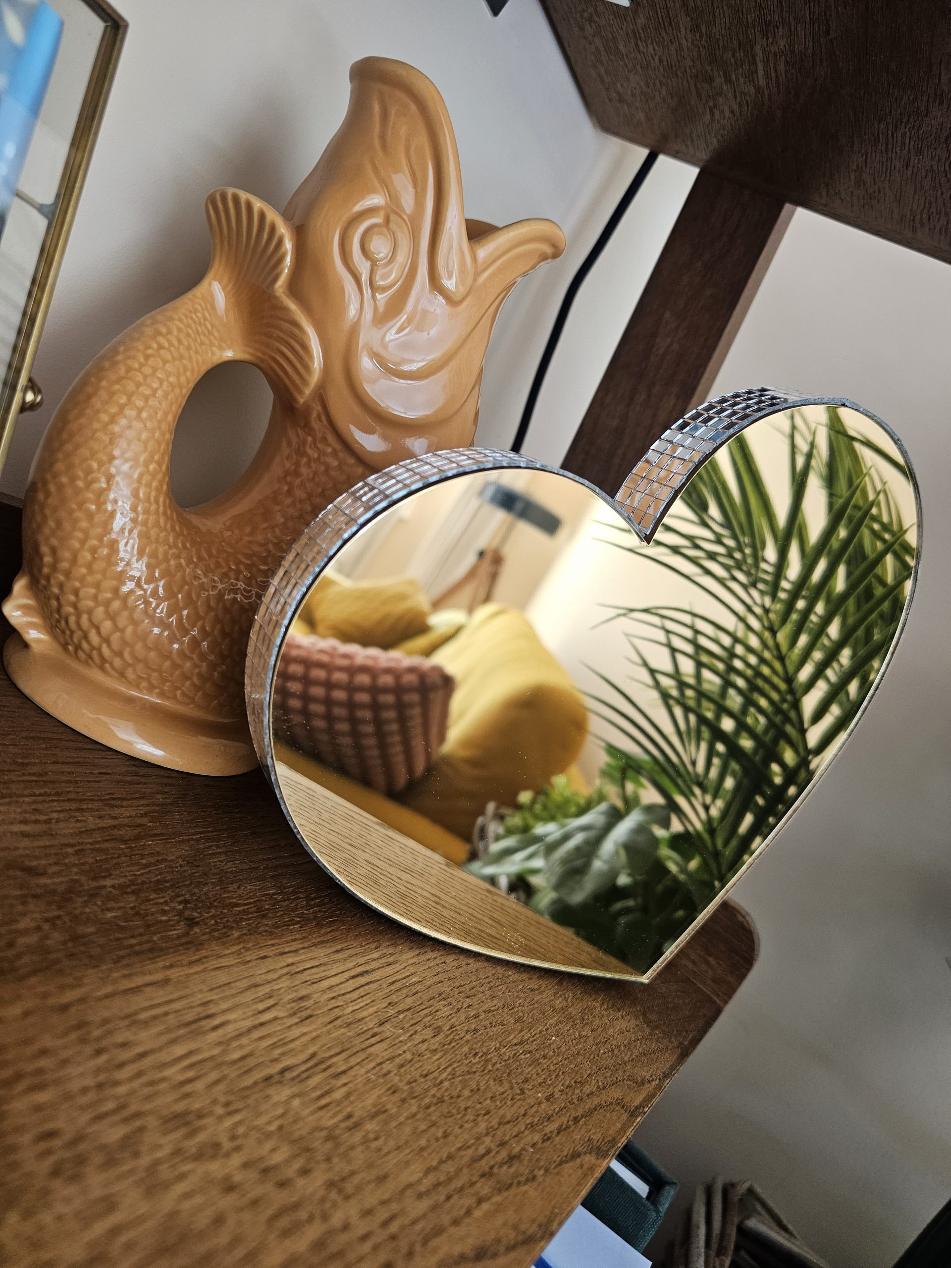 A gold mirror in the shape of a heart, with silver glass mirror tiles around the edges, perched on a shelf next to a vintage fish ornament.